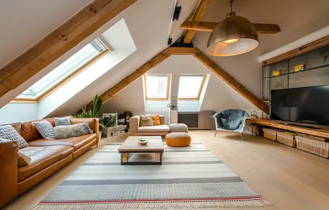A loft converted into a living room, with roof windows in the sloping ceiling, exposed timber beams, a pale wooden floor and a leather sofa