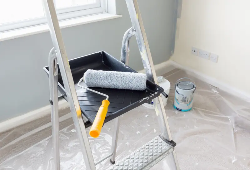A paint roller resting in a black tray on aluminium step ladders, in a room with dust sheets over the carpet and a wall part painted in grey